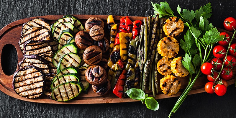 Auf einem großen Holzbrett liegen gegrillte Melanzani, Zucchini, Pilze, Paprika, Bohnen, Maiskolben und Rispentomaten. Daneben stehen ein Salz- und Pfefferstreuer.
