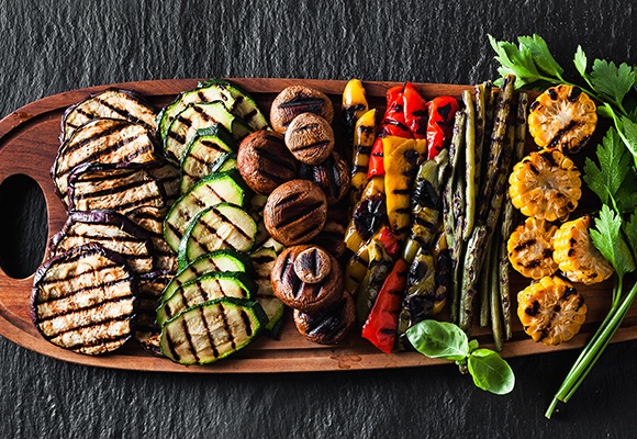 Auf einem großen Holzbrett liegen gegrillte Melanzani, Zucchini, Pilze, Paprika, Bohnen, Maiskolben und Rispentomaten. Daneben stehen ein Salz- und Pfefferstreuer.