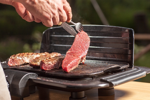 Ein Elektrogrill auf dem vier Steaks liegen. Eines wird von einem Mann mit Messer und Gabel gerade gewendet.