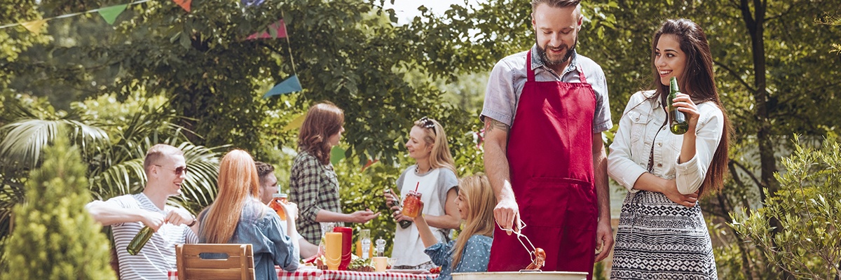 Freunde grillen gemeinsam im großen Garten. Der Grillmeister trägt eine rote Grillschürze und unterhält sich mit einer Frau, die ein Bier trinkt.