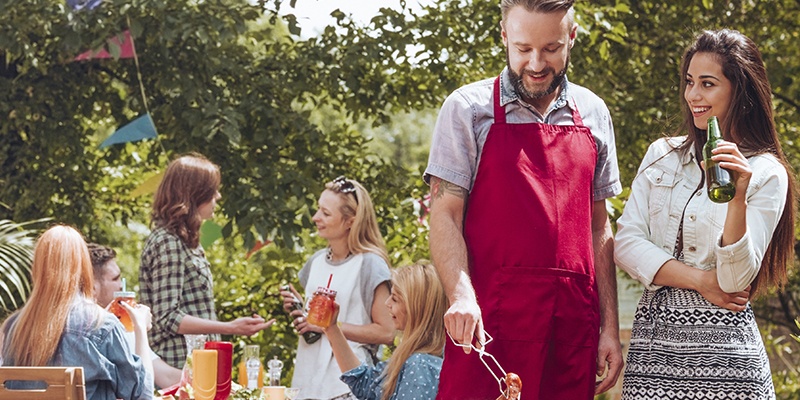 Freunde grillen gemeinsam im großen Garten. Der Grillmeister trägt eine rote Grillschürze und unterhält sich mit einer Frau, die ein Bier trinkt.