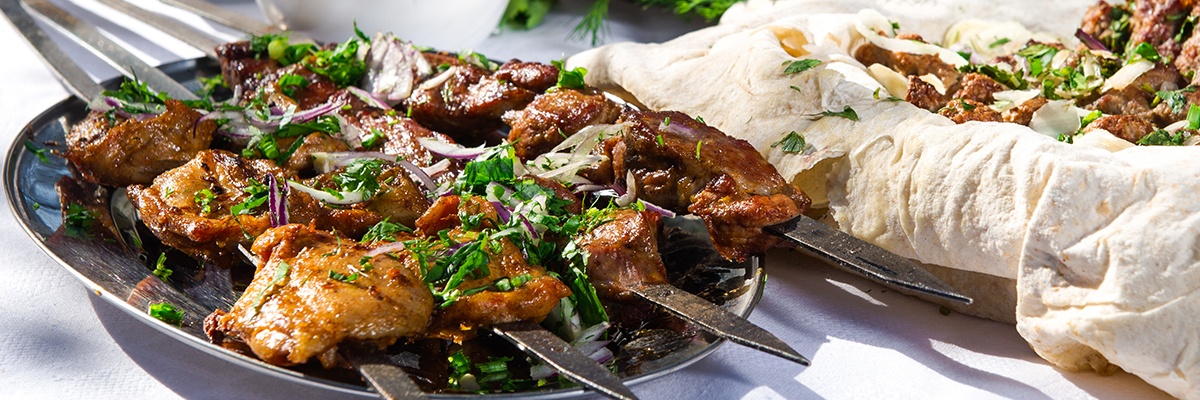 Grillfleisch ist auf einem Metallspieß aufgereiht und liegt auf einem silbernen Teller, daneben Wrapteig und weitere Spieße.