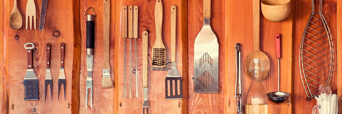 An einer Holzwand hängt unterschiedliches Grillzubehör, wie mehrere Fleischgabeln, ein Soßenpinsel, eine Grillbürste, Küchenhelfer, Grillzangen, ein Schöpflöffel und ein Fischbräter. Davor steht eine Glasflasche und ein Glas mit Plastikbesteck.