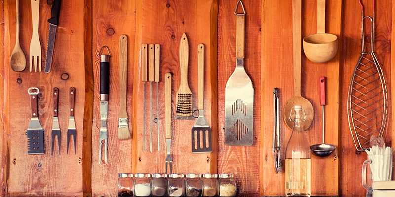 An einer Holzwand hängt unterschiedliches Grillzubehör, wie mehrere Fleischgabeln, ein Soßenpinsel, eine Grillbürste, Küchenhelfer, Grillzangen, ein Schöpflöffel und ein Fischbräter. Davor steht eine Glasflasche und ein Glas mit Plastikbesteck.
