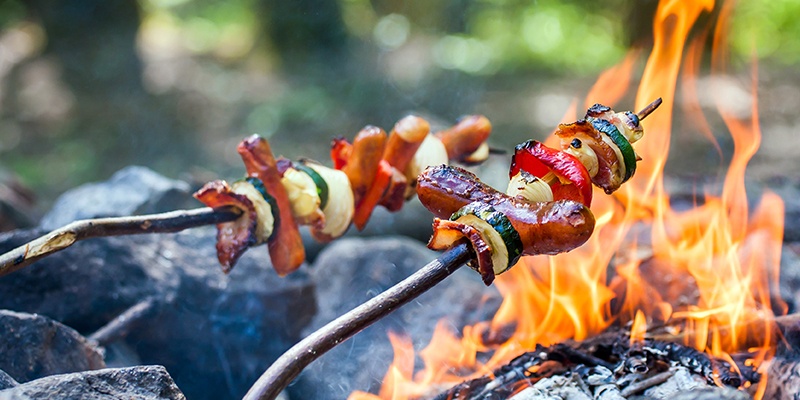 Grillspieße mit Würstchen, Paprika, Zucchini und Zwiebel auf einen Ast gereiht werden über einem Lagerfeuer gegrillt.