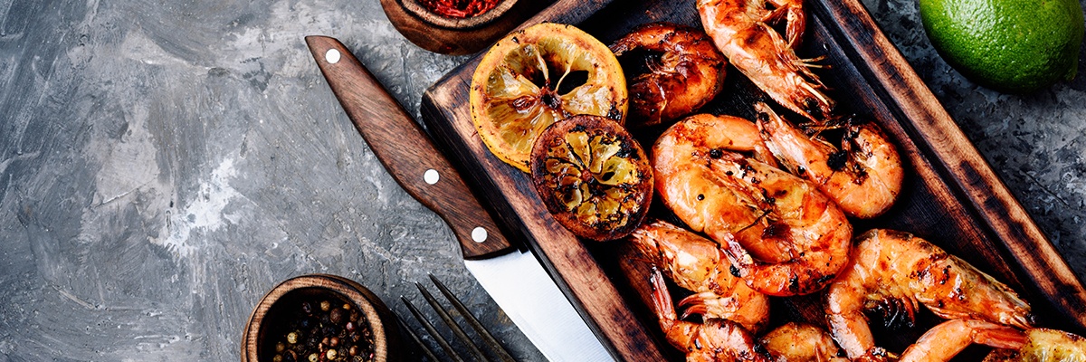 Garnelen liegen bräunlich gegrillt auf einem Holzbrettchen, daneben ein Messer und eine Gabel sowie Pfefferkörner.