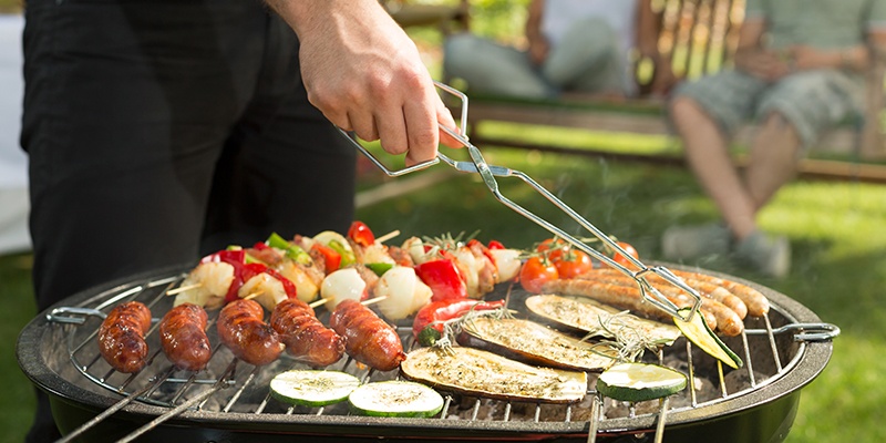 Auf einem runden Holzkohlegrill werden Gemüsespieße, unterschiedliche Bratwürste, Scheiben von Zucchini und Melanzani gegrillt. Mit einer Grillzange wird eine Zucchinischeibe gewendet.