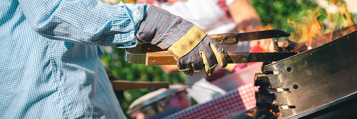 Ein Mann steht vor seinem Grill. Er trägt Schutzhandschuhe und hält eine Holzzange zum Wenden des Grillguts in seiner Hand.