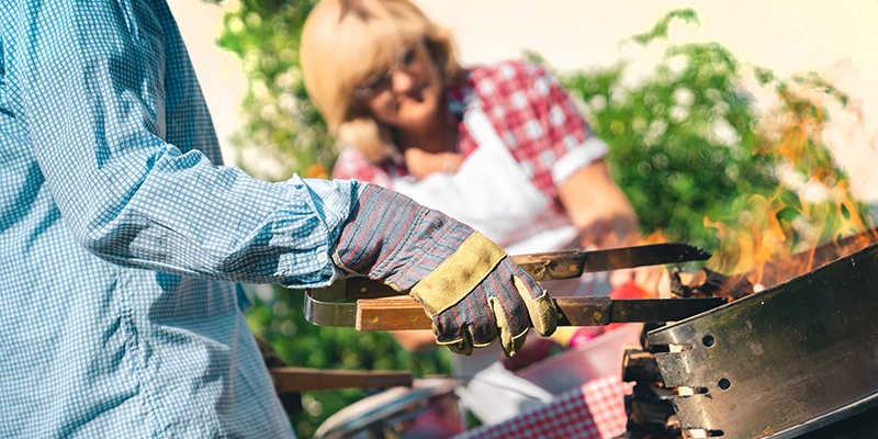Ein Mann steht vor seinem Grill. Er trägt Schutzhandschuhe und hält eine Holzzange zum Wenden des Grillguts in seiner Hand.