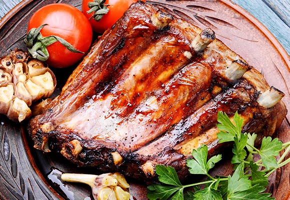 Spareribs mit Knoblauch auf einem Holzteller. Links daneben liegt ein Messer und eine Holzschale mit Gewürzen.
