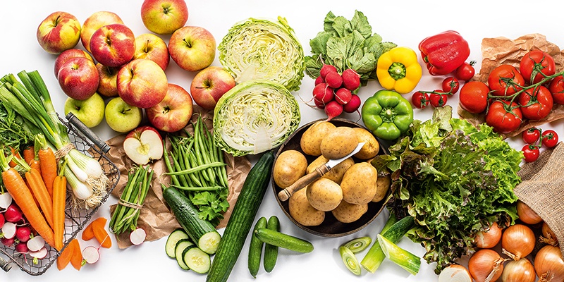 Auf weißem Hintergrund liegt unterschiedliches Obst und Gemüse. In der Mitte ist eine Schale mit Kartoffeln. 