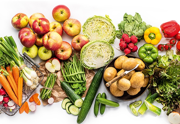 Auf weißem Hintergrund liegt unterschiedliches Obst und Gemüse. In der Mitte ist eine Schale mit Kartoffeln. 