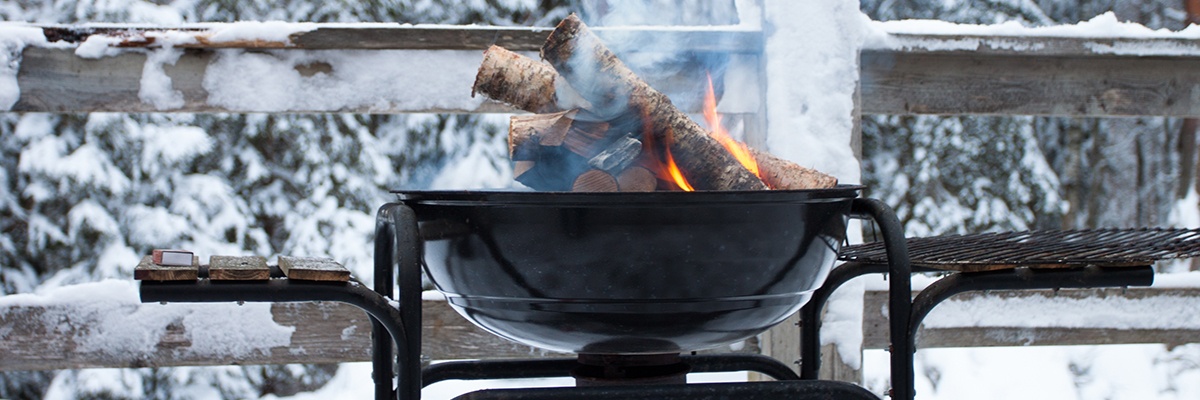 Vor einem eingeschneiten Wald und Zaun brennen Holzscheite in einer schwarzen Grillpfanne.