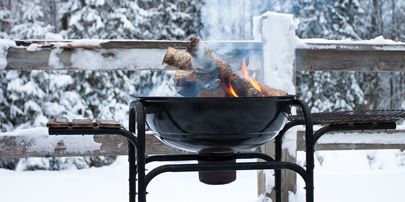 Vor einem eingeschneiten Wald und Zaun brennen Holzscheite in einer schwarzen Grillpfanne.