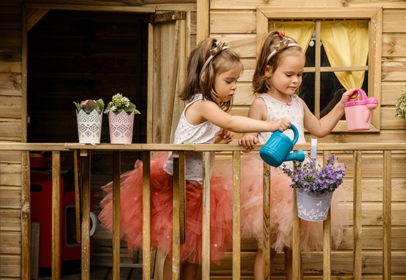 Zwei Mädchen stehen am Geländer ihres Baumhauses. Jede von ihnen hat eine kleine Gießkanne in der Hand. Das linke Mädchen gießt Blumen die an dem Geländer hängen.