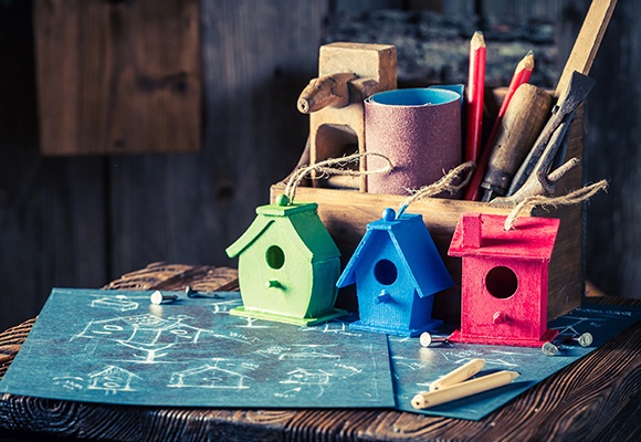 Ein grünes, ein blaues und ein rotes Vogelhäuschen stehen vor einer Holzkiste, die mit Werkzeug gefüllt ist.