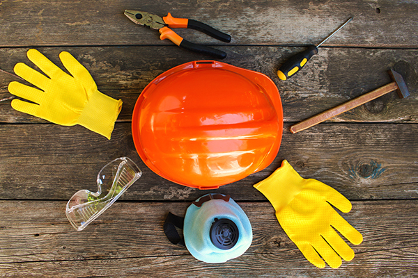 In der Mitte liegt ein oranger Bauarbeiterhelm. Kreisförmig darum angeordnet sind gelbe Sicherheitshandschuhe, eine Schutzbrille, eine Kombizange, ein Schraubenzieher, ein Hammer und ein Atemschutz.