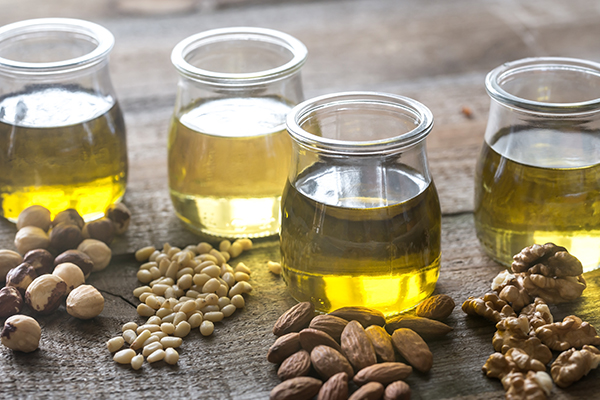 Vier Gläser mit unterschiedlichen pflanzlichen Ölen stehen auf einer Holzfläche. Davor sind unterschiedliche Nusssorten gestreut.