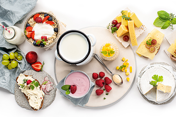 Joghurt und Topfen in Schüsseln von oben betrachtet. Daneben ist ein bestrichenes Brot, eine Müslischale, aufgeschnittener Hart- und Weichkäse.