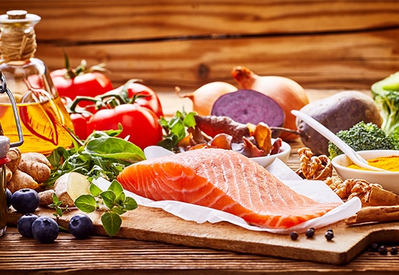 Ein Lachsfilet auf einem Holzbrett. Im Hintergrund ist Gemüse und eine Flasche mit Öl.