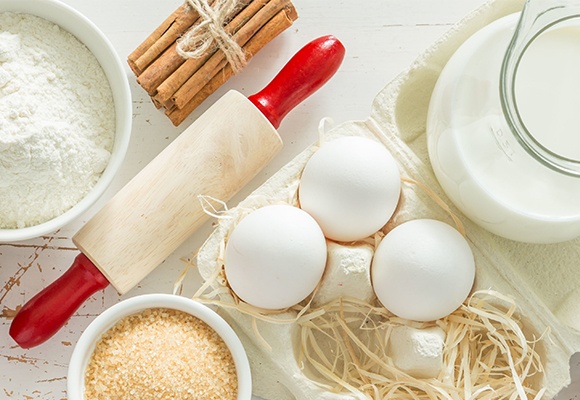 Eine Schüssel Mehl, eine mit braunem Zucker und drei weiße Eier. Daneben eine Kanne Milch und ein Bund Zimtstangen.