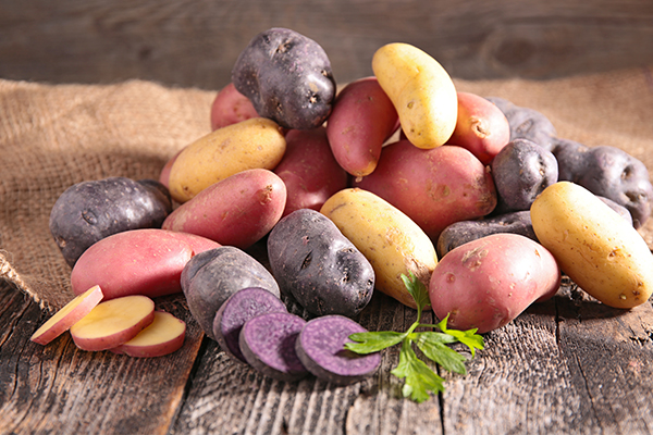 Unterschiedliche Kartoffelsorten liegen auf einem Holzuntergrund. Dahinter ein Jutesack.