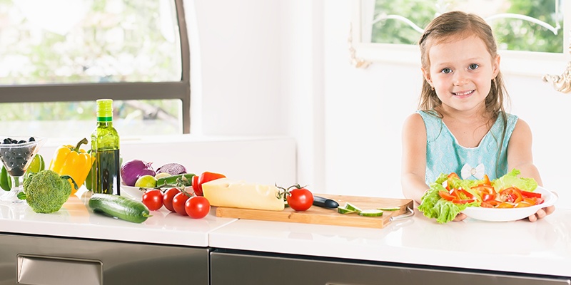 Ein lächelndes Mädchen stellt einen Teller mit geschnittenem Salat und Paprika auf einen Tisch.