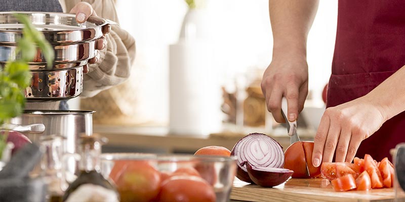 Ein Mann mit roter Schürze schneidet Tomaten. Vor im liegt eine in zwei Hälften geschnittene Zwiebel.