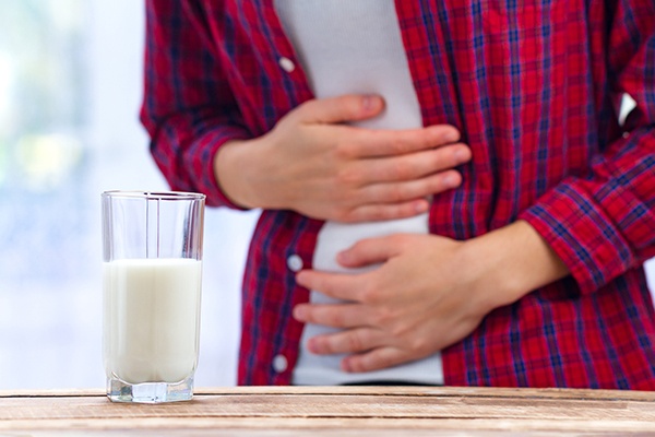 Ein Glas Milch steht im Vordergrund. Im Hintergrund sieht man einen Mann, der beide Hände auf den Bauch legt, als würde er schmerzen.