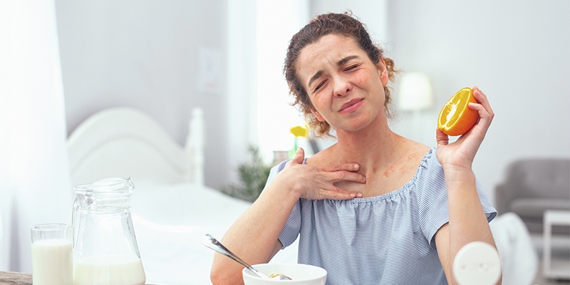 Eine Frau mit schmerzverzerrtem Gesicht hält eine halbe Orange in ihrer linken Hand. Mit ihrer rechten fasst sie sich an den Hals.