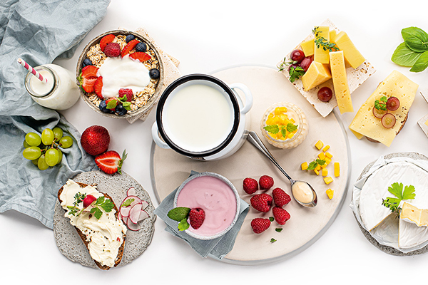 Joghurt und Topfen in Schüsseln von oben betrachtet. Daneben ist ein bestrichenes Brot, eine Müslischale, aufgeschnittener Hart- und Weichkäse.