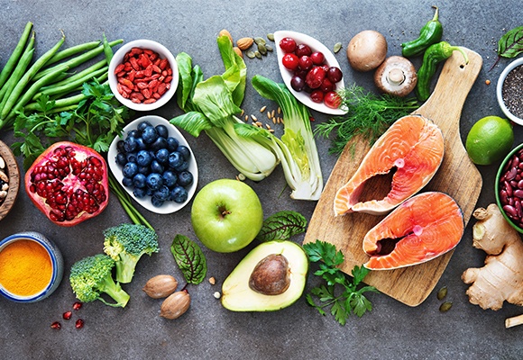 Ein aufgeschnittener Granatapfel, eine Schüssel mit Heidelbeeren, ein grüner Apfel, ein Holzschneidebrett mit aufgeschnittenem Fisch auf grauem Untergrund .