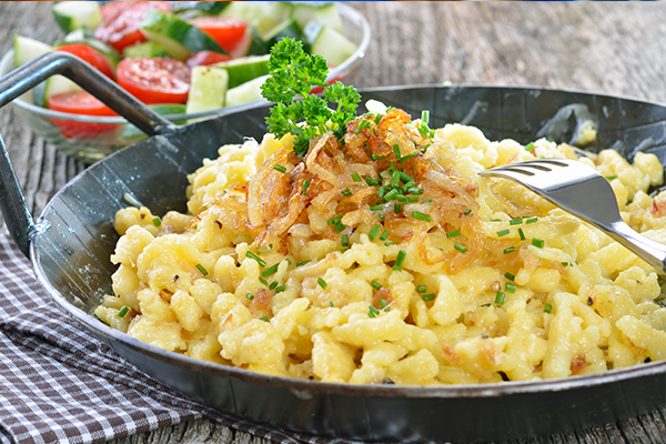 Käsespätzle in einer kleinen Metallpfanne, garniert mit Röstzwiebel und Petersilie. Dazu Gurken- und Tomatensalat.