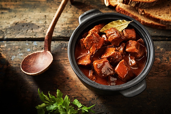 Ein Gulasch in einem kleinen Topf auf einem Untergrund aus Holz. Daneben ein hölzerner Schöpflöffel.