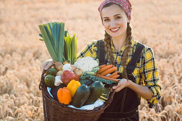 Eine Jungbäuerin mit zwei Zöpfen, die auf einem Getreidefeld steht. Sie hält einen Korb mit frischem Gemüse hoch.