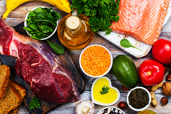 Auf einer Holzfläche liegen ein Stück Fleisch, mehrere Schüsseln gefüllt mit Linsen, Öl, Gewürzen und Salat. Auf einem Brett ist ein Lachsfilet. Daneben steht eine Flasche Öl