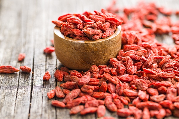 Eine kleine Holzschale mit roten Goji-Beeren. Daneben liegen noch weitere Beeren.