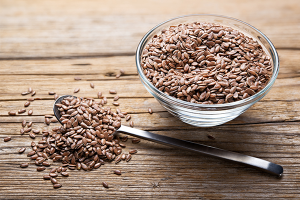 Eine kleine Glasschüssel gefüllt mit Leinsamen. Daneben ein mit Leinsamen gehäufter Teelöffel.