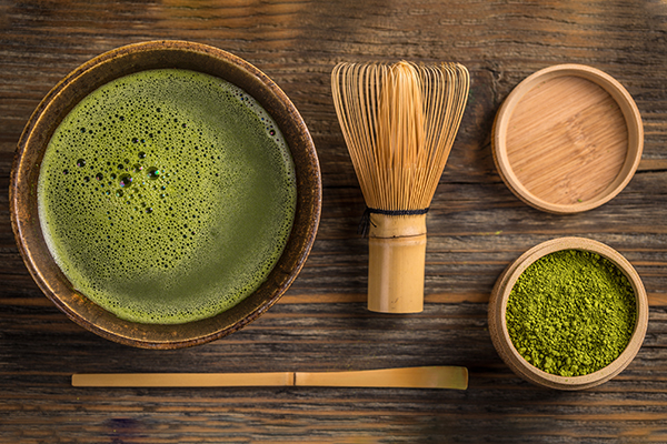 Eine Schale mit grünem Matcha, einem Bambusbesen, eine Holzdose mit grünem Matchapulver und einem Bambuslöffel liegen auf einer Holzfläche.