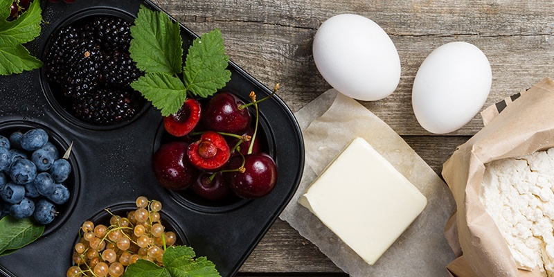 Backzutaten stehen auf einem rustikalen Tisch: Beeren wie Himbeeren, Heidelbeeren und Brombeeren sowie Eier, Butter und Mehl.