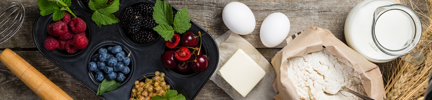 Backzutaten stehen auf einem rustikalen Tisch: Beeren wie Himbeeren, Heidelbeeren und Brombeeren sowie Eier, Butter, Mehl und Milch.