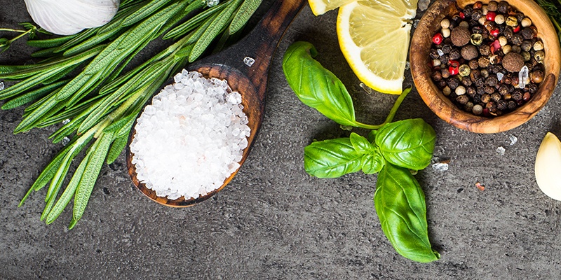 Kräuter, ein Holzlöffel mit groben Salz, Basilikum und ein Schälchen mit buntem Pfeffer auf grauem Hintergrund.