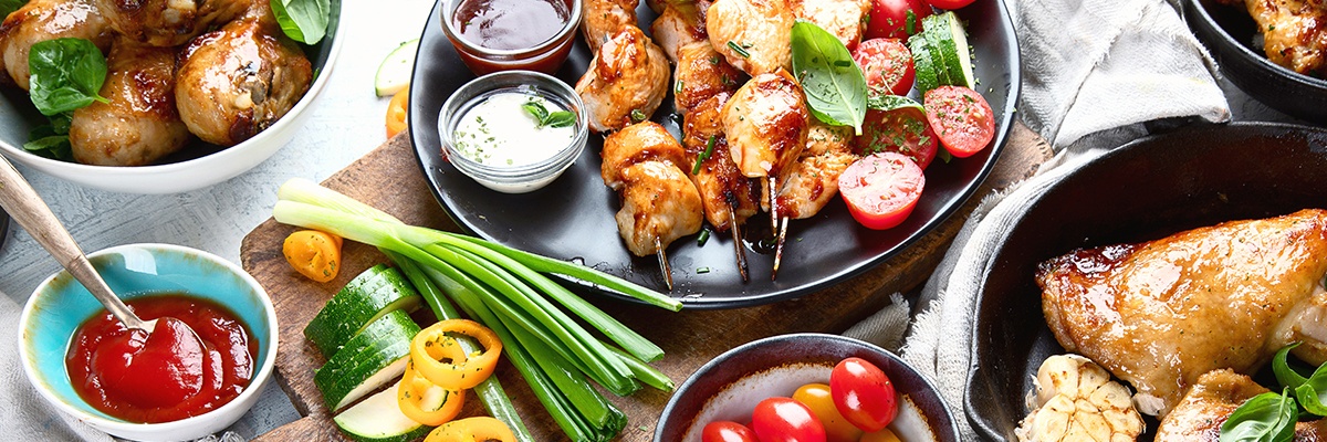 Zarte Hühnerspieße mit Dips und Frühlingszwiebeln sowie Tomaten liegen auf einem schwarzen Teller.