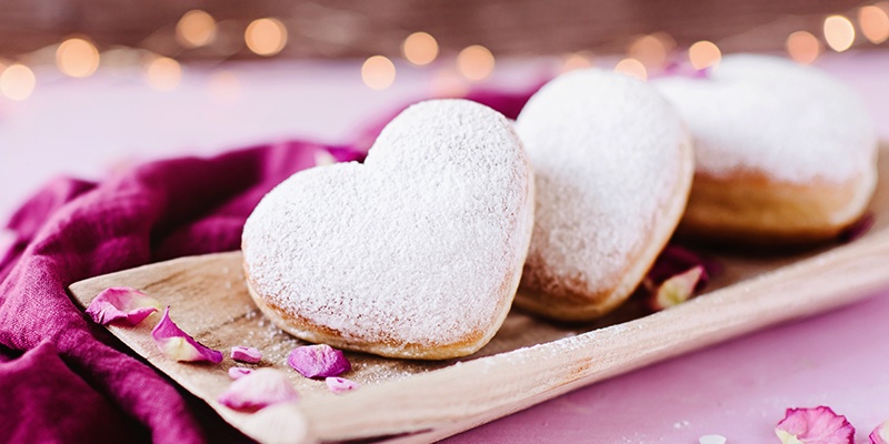 Pudrige Krapfen in Herzform liegen auf Holz in einem pinken Stoff.