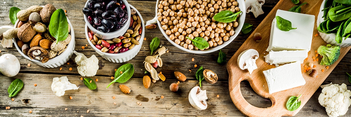 Nüsse aller Art in Schalen und grünes Gemüse, Pilze sowie Tofu liegen auf einem Holztisch.