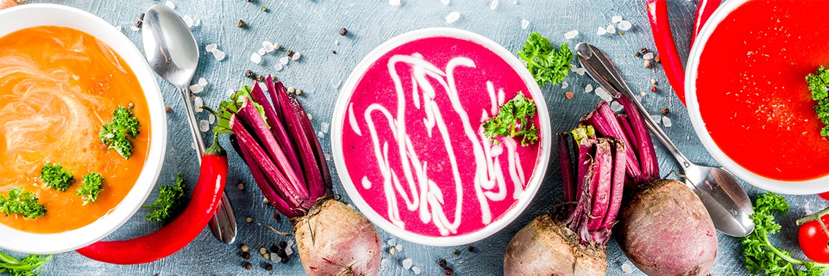 Drei Keramikschüsseln mit unterschiedlichen Suppen. In der linken ist eine Kürbiscremesuppe, in der Mitte eine Rote Bete Suppe und rechts eine rote Tomatensuppe. Verteilt liegen Zwiebel und eine Chili.