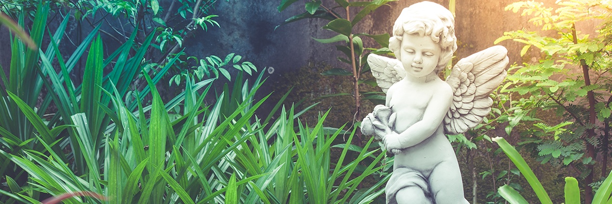 Eine Engelsputte sitzt auf einem Stein inmitten verschiedener Gräser und Pflanzen. Von rechts oben erstrahlt Licht.