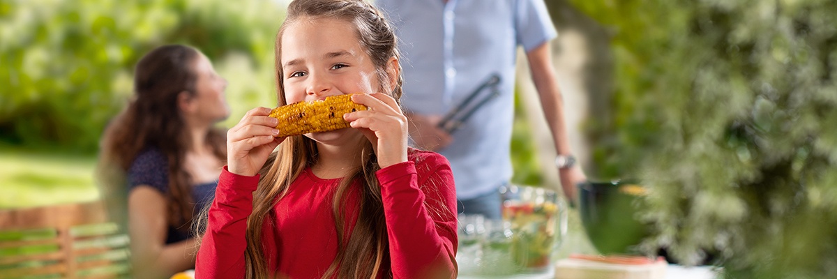 Foto zeigt ein Mädchen mit seinen Eltern im Garten, das einen Maiskolben isst.