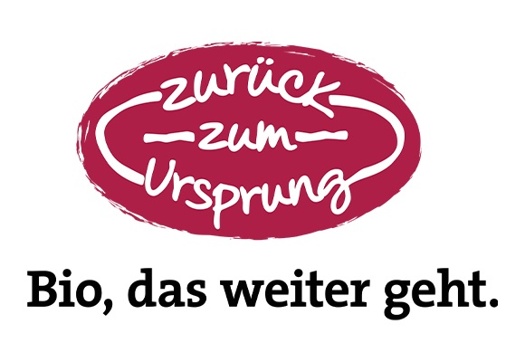 Ovales Logo mit dunkelrotem Rand, handgeschriebener weißer Schrift „zurück zum Ursprung“ und schwarzem Text darunter „Bio, das weiter geht.“