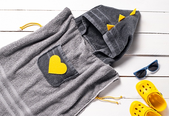 Graues Kapuzenhandtuch für kleine Kinder mit gelben Accessoires und Sonnenbrille auf einem Holzsteg